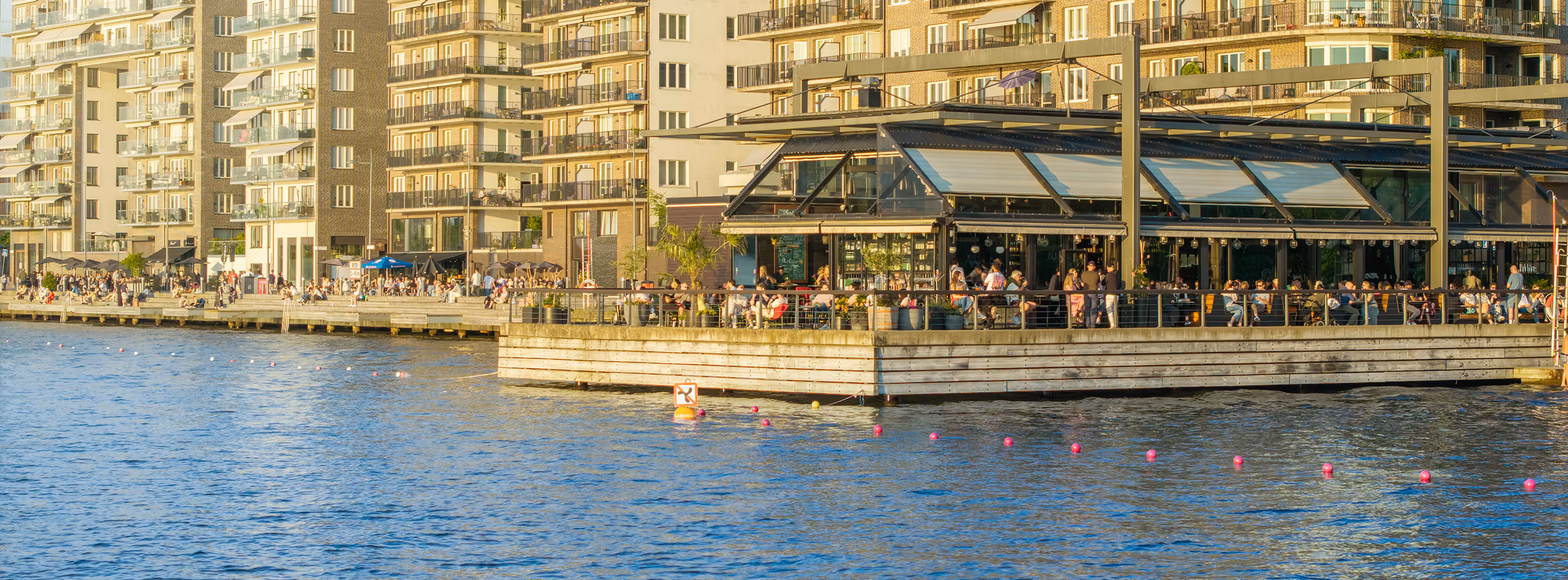 Piren sedd från vattnet: den glasade uteserveringen på bryggan i Hornsbergs Strand, full av gäster en solig sommarkväll.